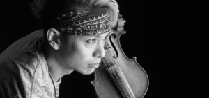 Black and white photograph of KATEI holding a violin. He is facing towards the right of the image.