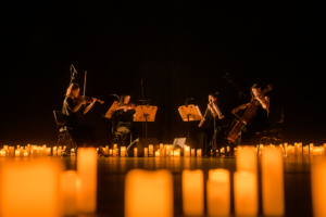 Photo from a previous Candlelight Concert. A string quartet is surrounded by candles on a dimly lit stage.