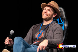 Liam McIntyre on stage at Supanova 2021 - Gold Coast. He is wearing a brown, brimmed hat, a dark T-shirt with a graphic, blue jeans and a grey sweater.