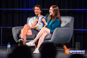 Bob Morley and Eliza Taylor on stage at Supanova 2022 - Adelaide. Eliza is wearing a teal blazer and white running shoes, while Bob is in shorts and a grey sweater. They are sitting on a couch laughing, talking to the audience.