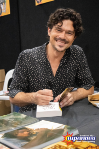 Author Luke Arnold signing autographs at Supanova 2021 - Brisbane. He is smiling at the camera, wearing a black and white spotted button-up shirt. He is signing a book in front of various headshots of his onscreen work.