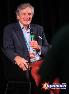 A photo of Nicholas Hammond smiling, holding a microphone on-stage, at Supanova 2022 - Melbourne.