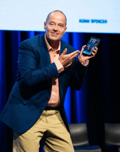 Adam Spencer on stage gesturing at a smartphone.